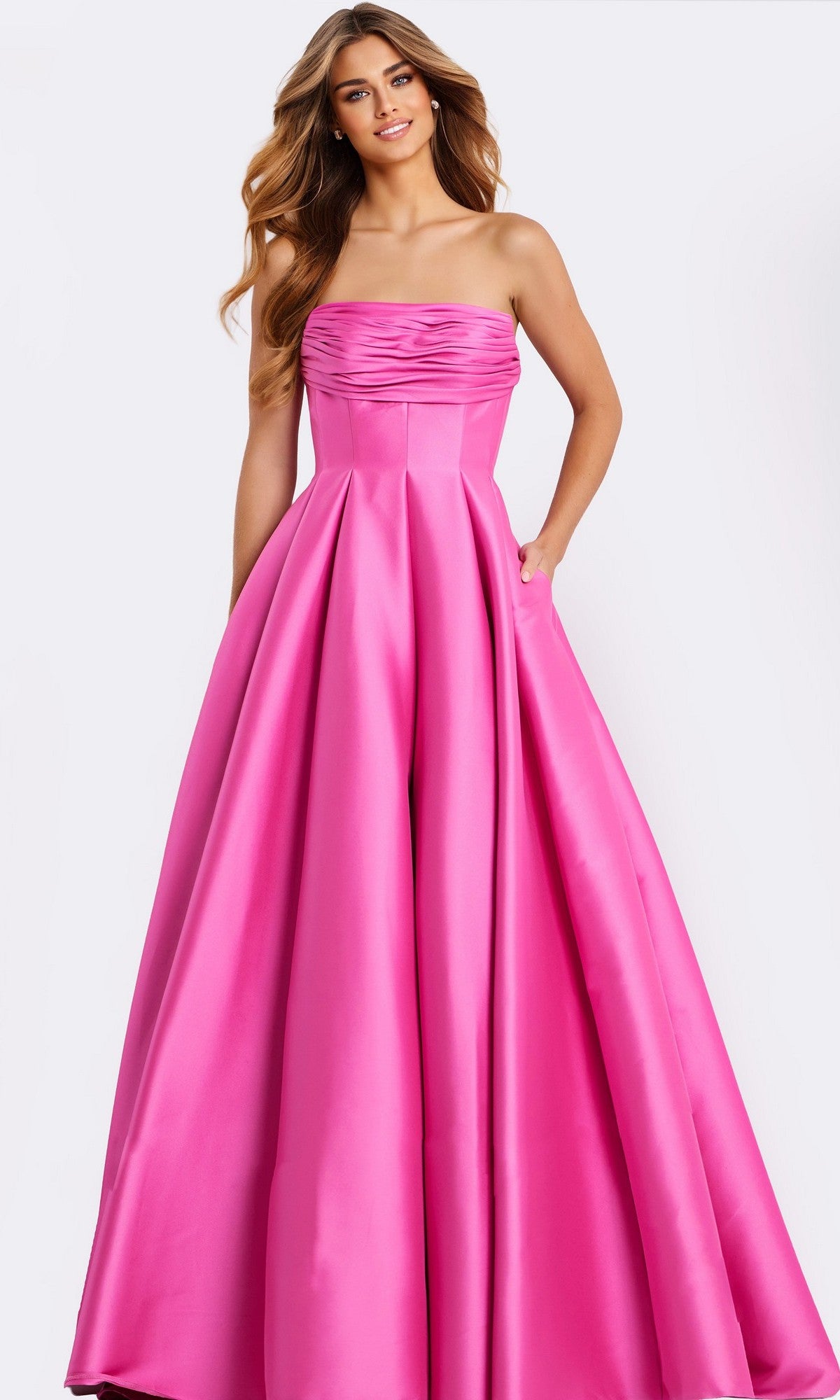 A woman wears the Strapless Long Pleated Prom Dress: Jovani 47570 in bright pink satin, featuring a fitted bodice and floor-length A-line skirt, standing against a plain light background.